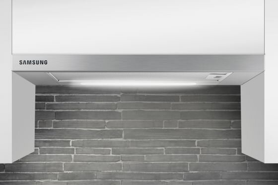 Okap kuchenny w kolorze stali nierdzewnej z widocznym logo \'SAMSUNG\'. Tło stanowi ściana z szarych cegiełek.