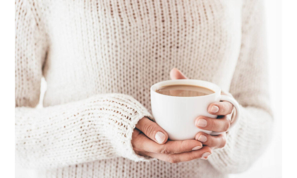 Osoba w jasnym swetrze trzyma w dłoniach białą filiżankę z kawą.