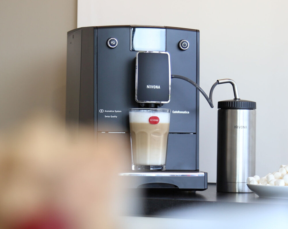 Ekspres do kawy marki NIVONA z napisem \'CafeRomatica\' przygotowuje kawę w szklance. Obok ekspresu stoi metalowy pojemnik z napisem \'NIVONA\'.