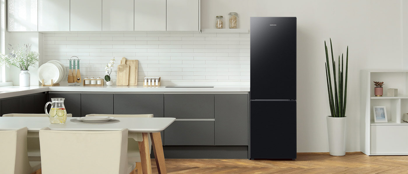 Nowoczesna kuchnia z czarną lodówką marki Samsung, umieszczoną obok wysokiej rośliny w doniczce. Wnętrze jest jasne, z białymi szafkami i drewnianą podłogą.