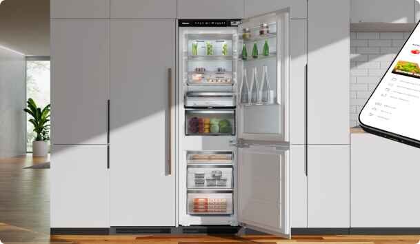 Otwarta lodówka w zabudowie kuchennej z widocznymi półkami wypełnionymi produktami spożywczymi, takimi jak butelki, słoiki i warzywa. Obok lodówki widoczny jest fragment smartfona z aplikacją na ekranie.