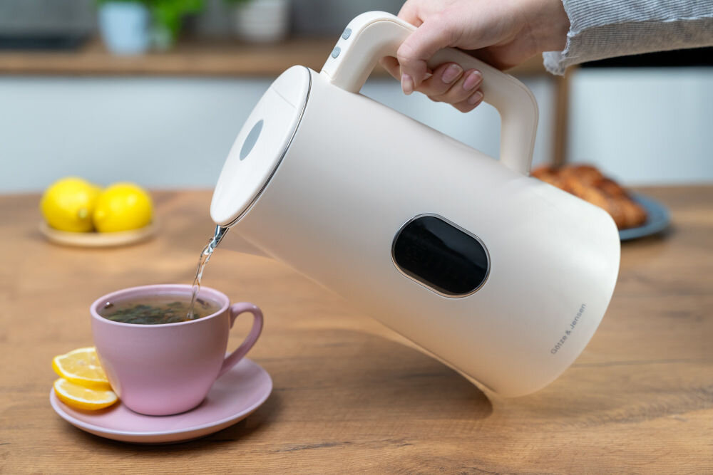 Osoba nalewa wodę z ecru czajnika do różowej filiżanki stojącej na drewnianym stole. Na czajniku widoczne jest logo \'GÖTZE & JENSEN\'.