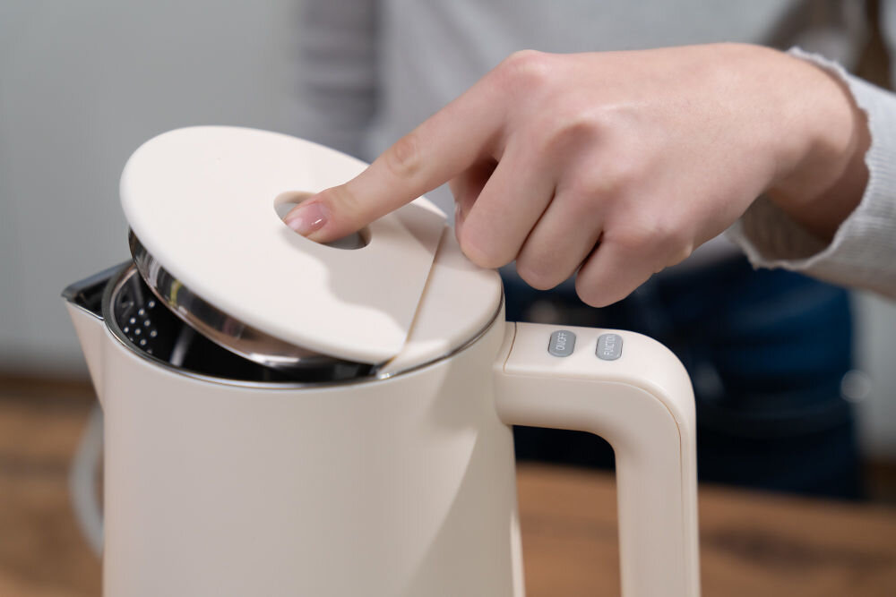 Osoba otwiera pokrywę czajnika w kolorze ecru, na uchwycie widoczne są przyciski z napisem \'OFF\'.