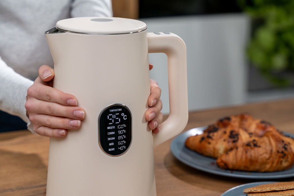 Osoba trzyma czajnik w kolorze ecru z wyświetlaczem pokazującym temperaturę 25°C. W tle na stole znajduje się talerz z croissantem.