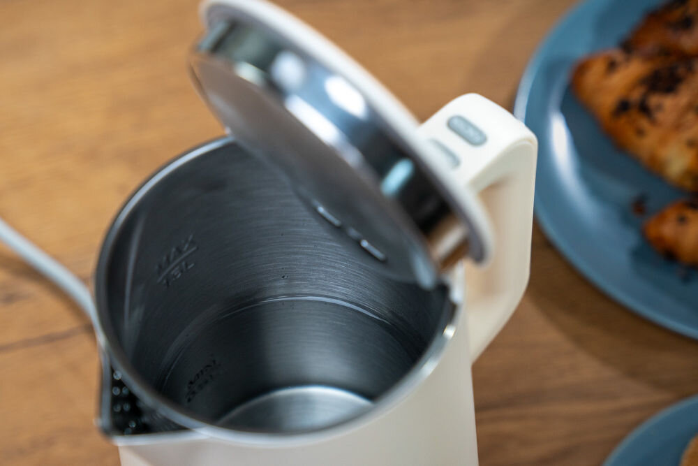 Otwarty czajnik z widocznym wnętrzem ze stali nierdzewnej, na którym znajduje się oznaczenie \'MAX 1.7L\'. Obok czajnika na stole leży talerz z rogalikami.