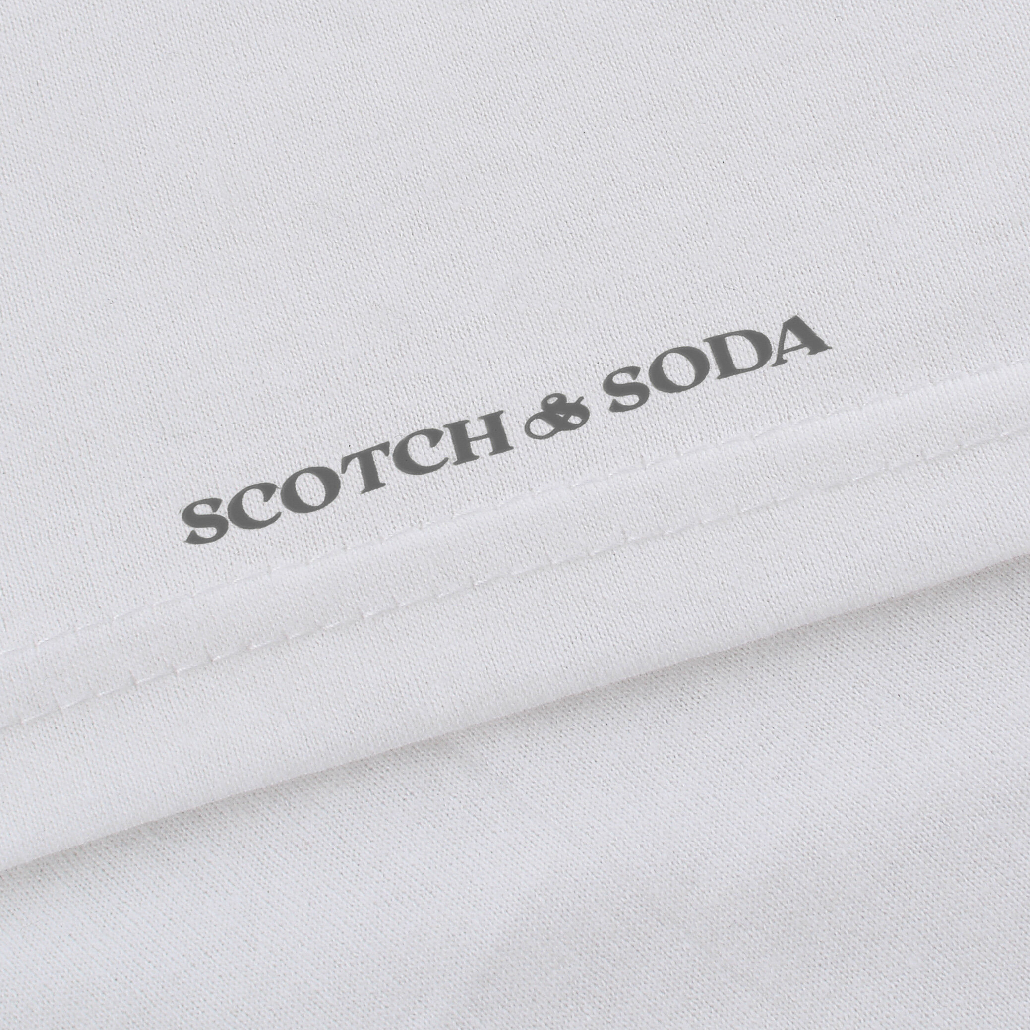 Biała tkanina z widocznym logo \'SCOTCH & SODA\' nadrukowanym w ciemnym kolorze przy krawędzi materiału.