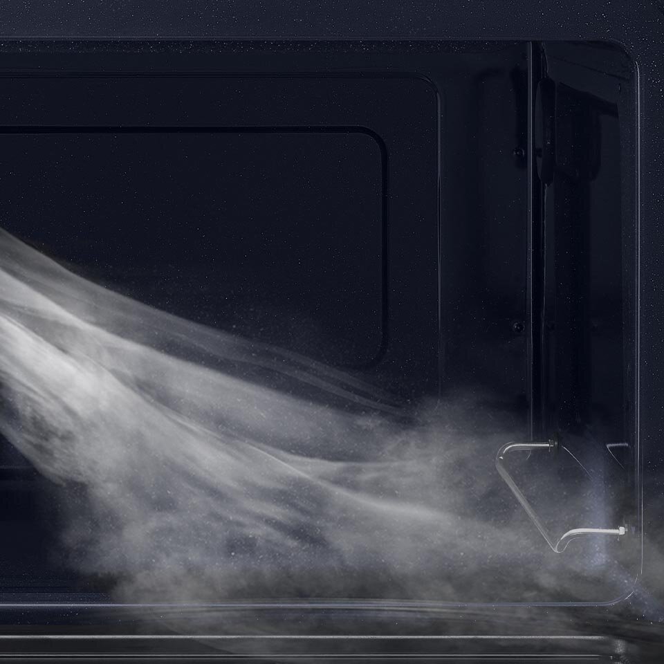 Wnętrze kuchenki mikrofalowej z widoczną parą unoszącą się wewnątrz. Ściany urządzenia są ciemne, a na dole widoczna jest metalowa kratka.