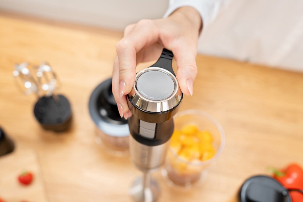 Osoba trzyma blender ręczny z metalowym wykończeniem, obok znajdują się pojemniki z pokrojonymi owocami i akcesoria kuchenne na drewnianym blacie.