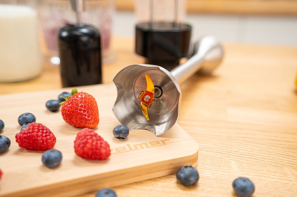 Na drewnianej desce z logo \'zelmer\' leżą świeże owoce: truskawki, maliny i jagody. Obok znajduje się metalowa końcówka blendera z widocznymi ostrzami.