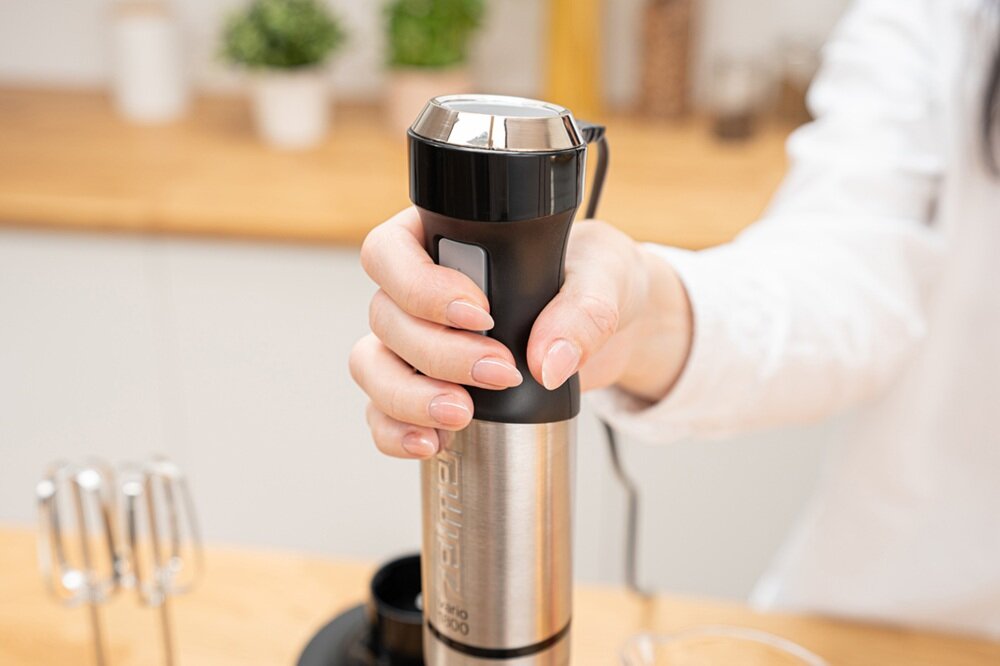 Osoba trzyma blender ręczny z metalową obudową i czarnymi elementami. W tle widać rozmytą kuchnię z roślinami w doniczkach.