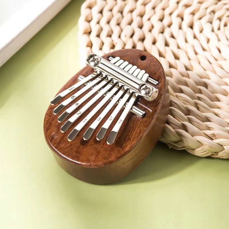 Na obrazie widoczna jest mini kalimba wykonana z drewna mahoniowego, z metalowymi klawiszami oznaczonymi numerami. Instrument leży na plecionym podkładzie na jasnozielonym tle.