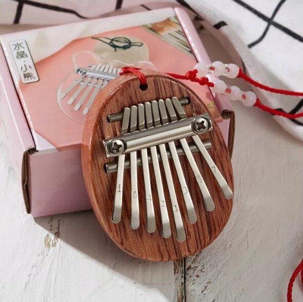 Na obrazie widoczna jest mini kalimba wykonana z drewna mahoniowego z metalowymi klawiszami oznaczonymi cyframi. W tle znajduje się różowe pudełko z grafiką kalimby i chińskimi znakami.