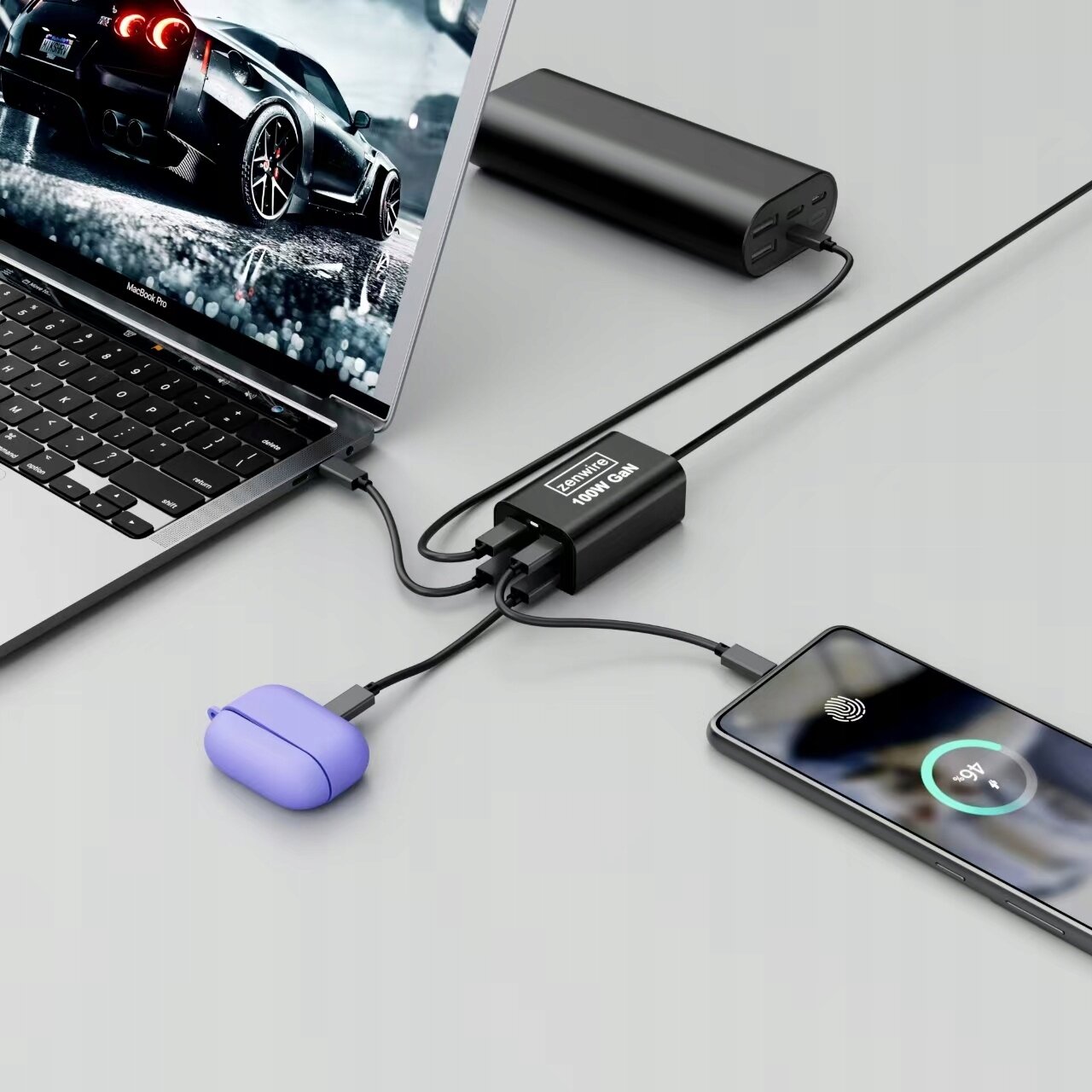 Na obrazie widać laptopa, telefon i słuchawki podłączone do czarnej ładowarki z napisem \'Zenwire 100W GaN\'. Obok znajduje się czarny powerbank.