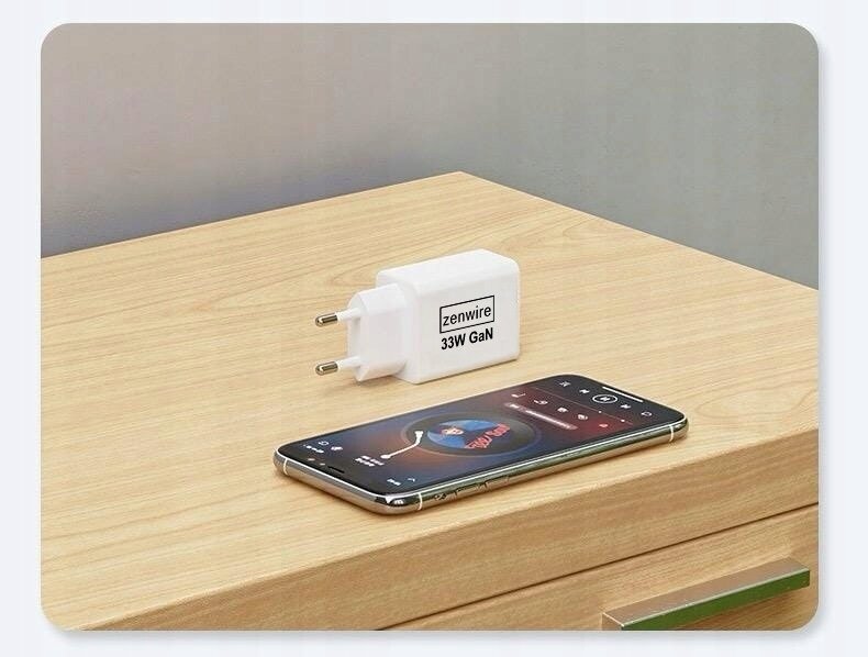 Na drewnianym blacie leży biały zasilacz z napisem \'zenwire 33W GaN\' oraz smartfon z wyświetlonym ekranem odtwarzacza muzyki.