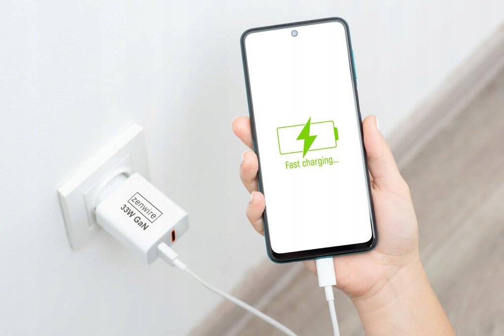 Osoba trzyma telefon z wyświetlonym napisem \'Fast charging...\' i ikoną baterii. Telefon jest podłączony do ładowarki w gniazdku z napisem \'Zenwire 33W GAN\'.