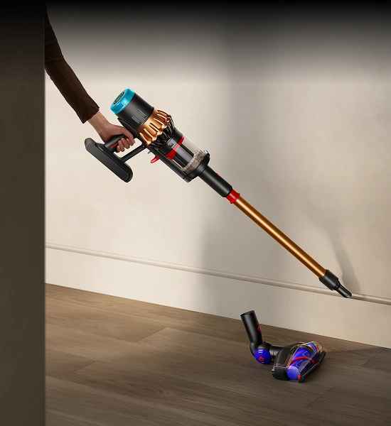 Osoba trzyma odkurzacz pionowy z długą, złotą rurą i przezroczystym pojemnikiem na kurz. Odkurzacz jest używany na drewnianej podłodze przy ścianie.