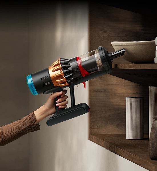 Osoba trzyma odkurzacz ręczny z przezroczystym pojemnikiem na kurz i kolorowymi elementami, używając go do czyszczenia półki. W tle widoczne są drewniane półki z dekoracyjnymi przedmiotami.