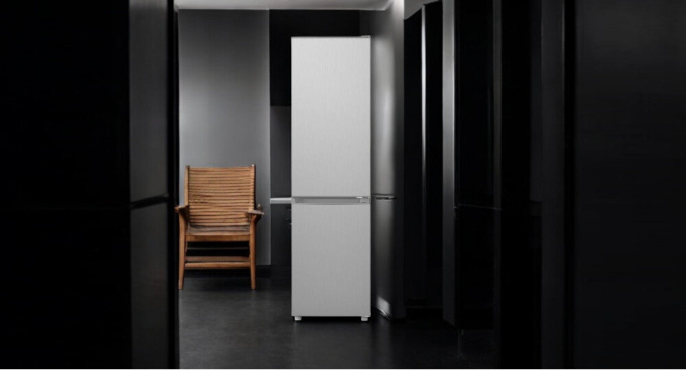 Na obrazie widoczna jest lodówka w kolorze inox, ustawiona w minimalistycznym wnętrzu z ciemnymi ścianami. Obok lodówki znajduje się drewniane krzesło.