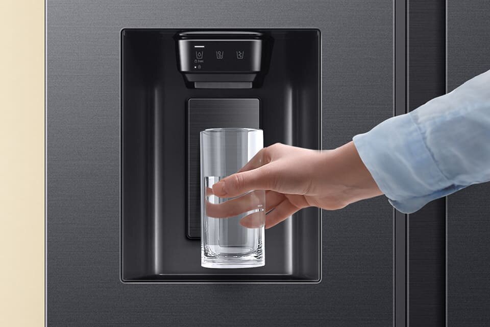 Osoba trzyma szklankę pod dozownikiem wody w lodówce. Na panelu widoczne są ikony z symbolami wody i kostek lodu.