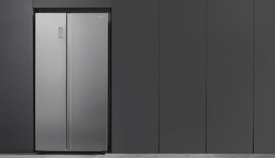 Srebrna lodówka side by side z panelem sterowania na drzwiach. W tle ciemne, gładkie panele ścienne.