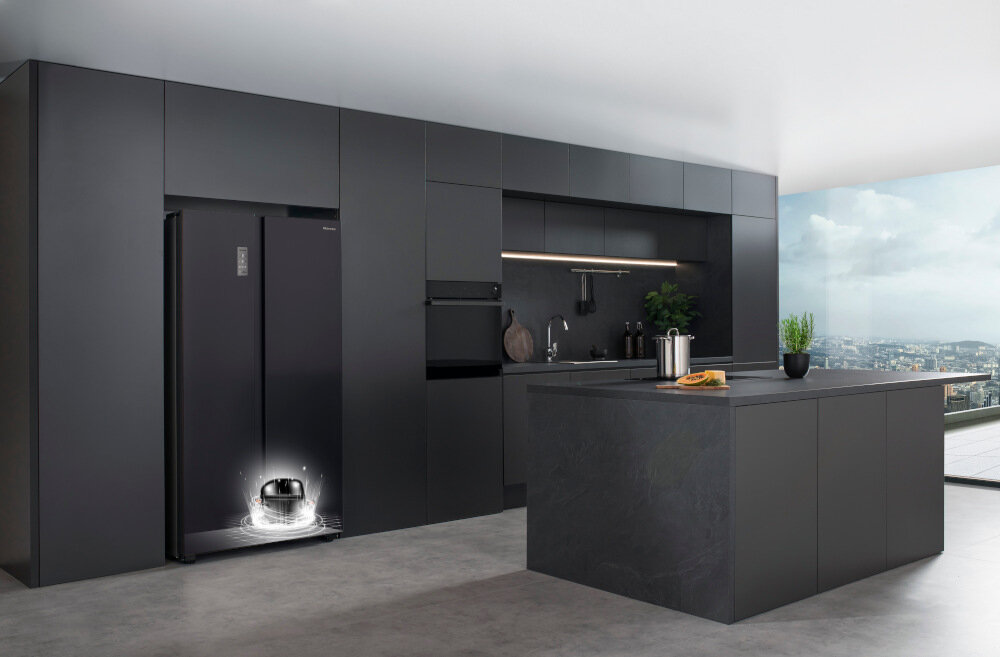 Czarna lodówka w zabudowie kuchennej z widocznym logo Hisense. Na blacie wyspy kuchennej stoi deska z jedzeniem i metalowy pojemnik.
