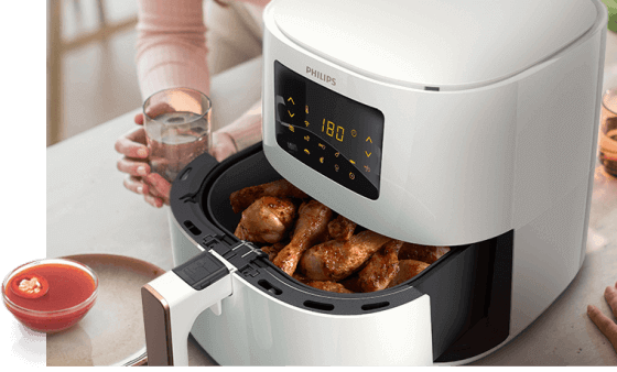Biała frytkownica beztłuszczowa z wyświetlaczem pokazującym temperaturę 180 stopni, wypełniona kawałkami kurczaka. Obok znajduje się osoba trzymająca szklankę z napojem oraz miseczka z sosem.