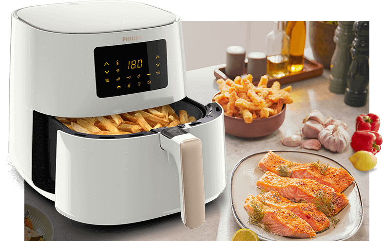 Na blacie kuchennym stoi biała frytkownica beztłuszczowa z otwartą szufladą pełną frytek. Obok znajduje się talerz z kawałkami łososia oraz miska z frytkami, a w tle widoczne są przyprawy i warzywa.