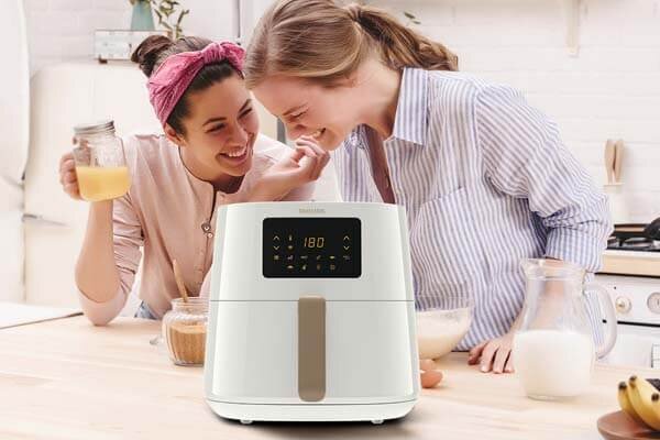 Na blacie kuchennym stoi biała frytkownica beztłuszczowa z wyświetlaczem pokazującym \'180\'. W tle dwie osoby przygotowują posiłek, otoczone różnymi naczyniami i składnikami.