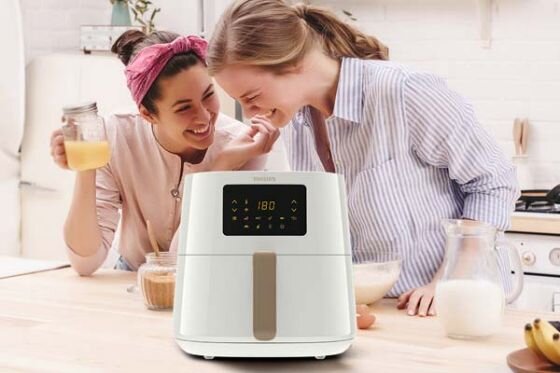 Na blacie kuchennym stoi biała frytkownica beztłuszczowa z wyświetlaczem pokazującym \'180\'. W tle dwie osoby przygotowują posiłek, jedna trzyma szklankę z sokiem pomarańczowym.