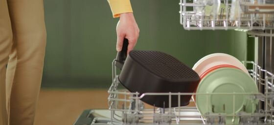 Osoba wkłada czarny koszyk do zmywarki, obok znajdują się kolorowe talerze. Koszyk ma perforowaną powierzchnię.