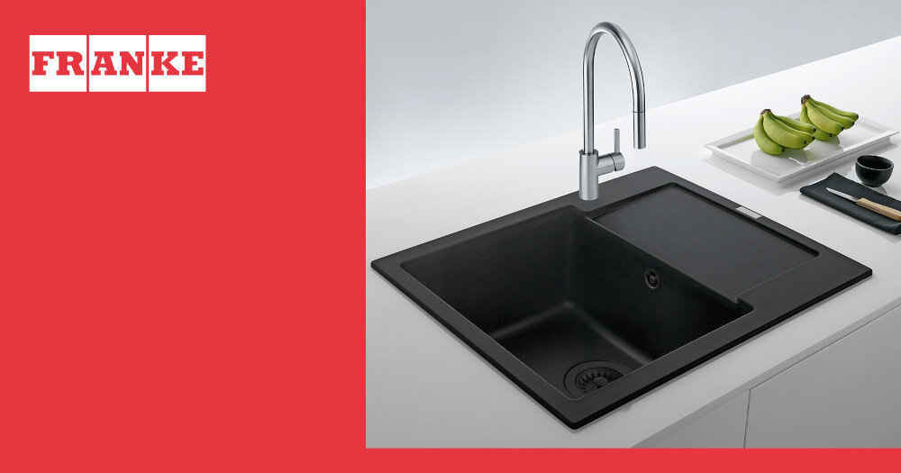 Czarny matowy zlewozmywak jednokomorowy z ociekaczem i srebrnym kranem, umieszczony na białym blacie. W tle widoczne są zielone banany i sztućce na czarnym talerzu, a po lewej stronie czerwone tło z białym logo \'FRANKE\'.