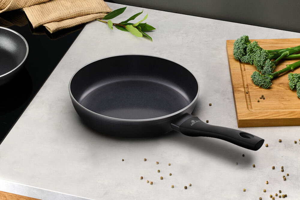 Na blacie kuchennym znajduje się czarna patelnia z uchwytem, obok której leży deska do krojenia z brokułami. W tle widoczne są zielone liście i rozsypane ziarna pieprzu.