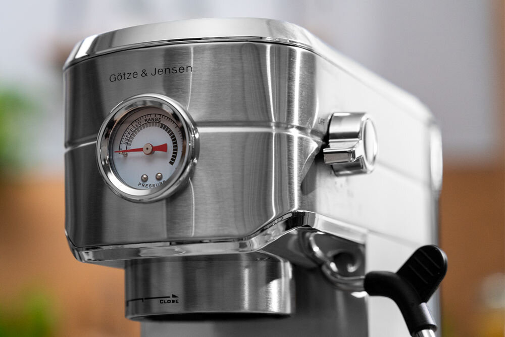 Ekspres do kawy wykonany ze stali nierdzewnej z widocznym logo \'Götze & Jensen\'. Na urządzeniu znajduje się okrągły wskaźnik ciśnienia z czerwoną wskazówką oraz pokrętło.