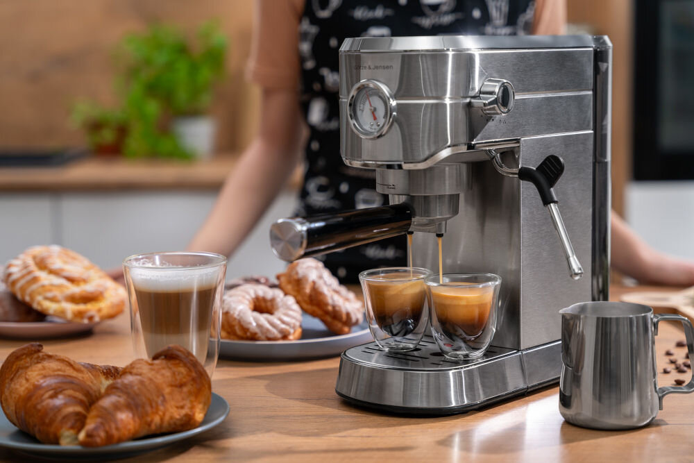 Ekspres do kawy ze stali nierdzewnej z logo \'GÖTZE & JENSEN\' przygotowuje dwa espresso. W tle widoczne są talerze z rogalikami i ciastkami oraz szklanka z kawą.