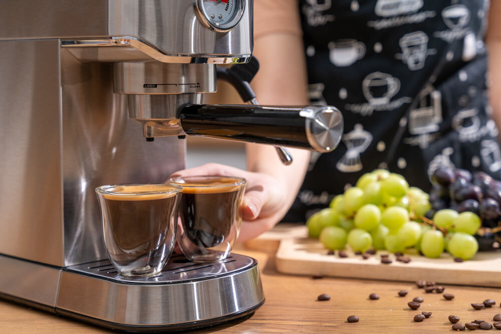 Ekspres do kawy z metaliczną obudową przygotowuje dwie filiżanki espresso. Obok znajduje się deska z zielonymi winogronami i rozsypanymi ziarnami kawy.