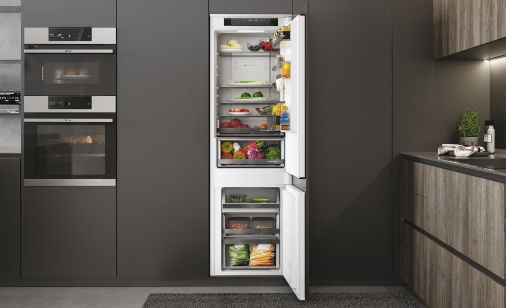 Wnętrze otwartej lodówki w zabudowie, wypełnionej różnorodnymi produktami spożywczymi. Obok znajdują się zabudowane piekarniki i szafki kuchenne.