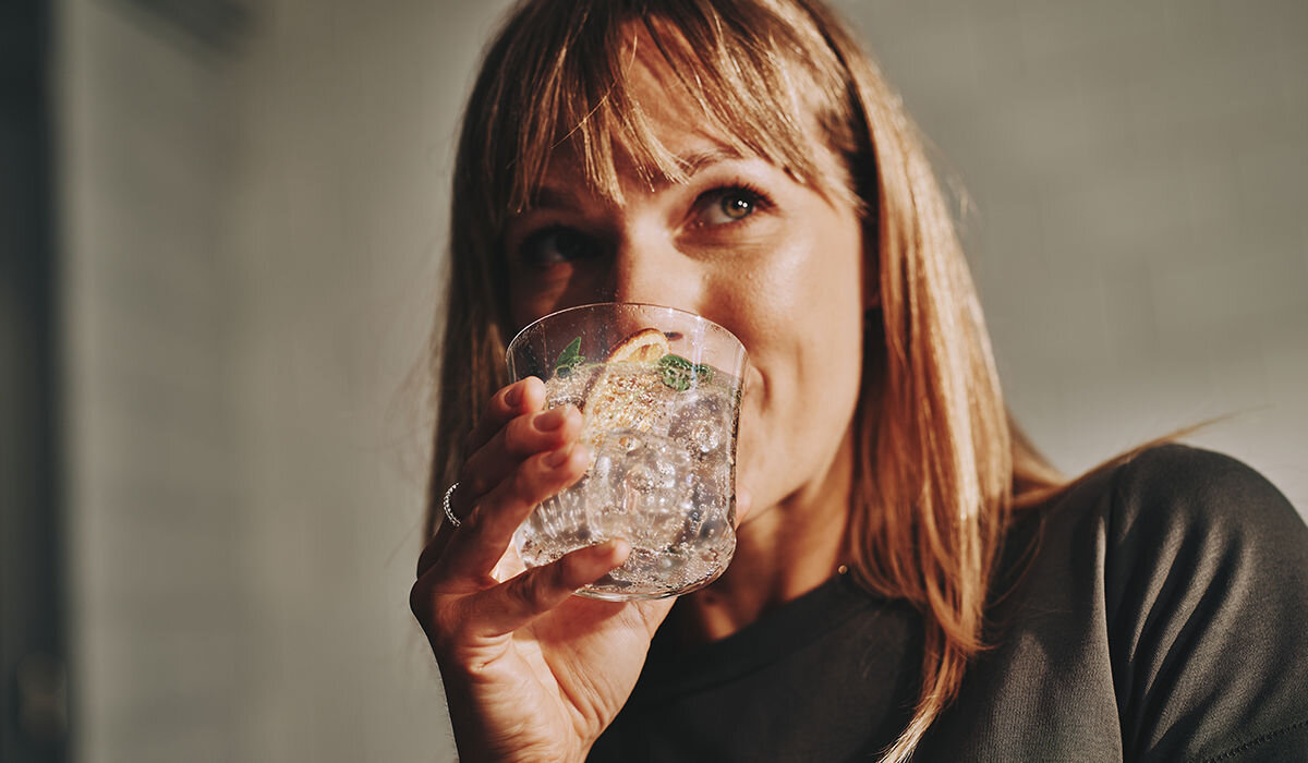 Osoba trzyma szklankę z napojem gazowanym, w której znajdują się plasterki cytryny i liście mięty.