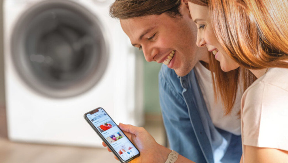 Dwie osoby patrzą na smartfon, na którym widoczna jest aplikacja sklepu media expert. W tle znajduje się urządzenie przypominające pralkę lub suszarkę.