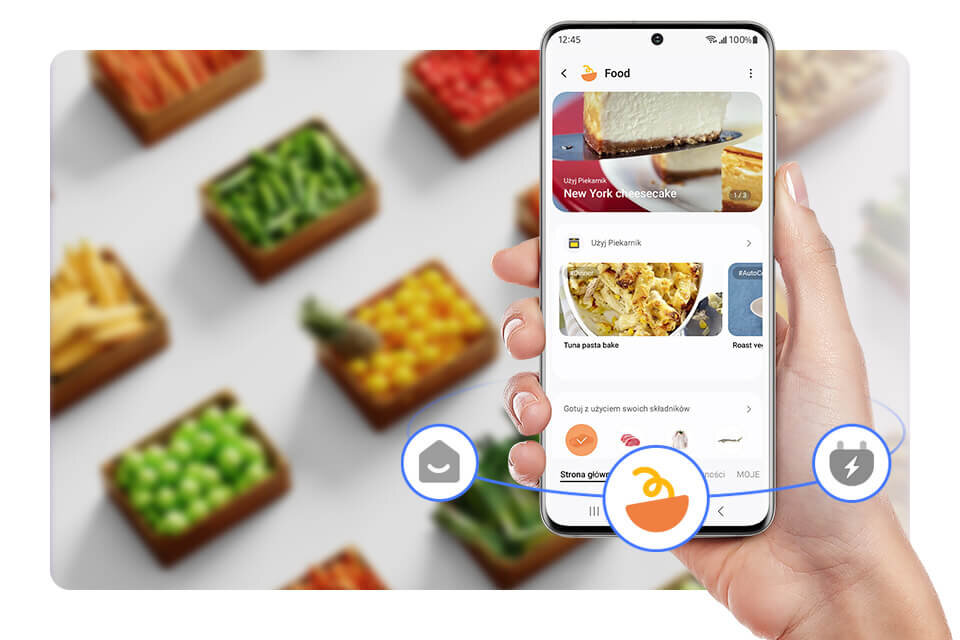 Osoba trzyma smartfon z aplikacją kulinarną, na ekranie widoczny jest przepis na \'New York Cheesecake\'. W tle znajdują się pudełka z różnokolorowymi warzywami.