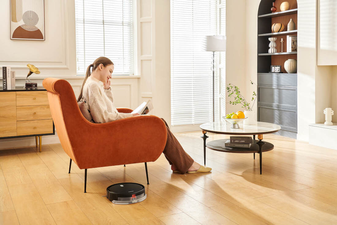 W jasnym pokoju na drewnianej podłodze znajduje się czarny robot sprzątający. Obok siedzi osoba na pomarańczowym fotelu, trzymająca książkę.