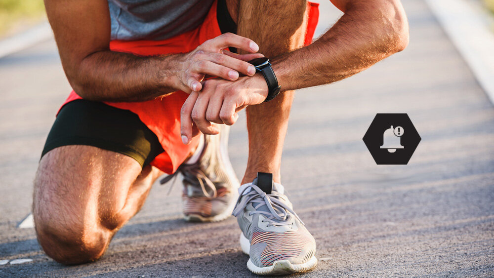 Osoba w sportowym stroju klęczy na asfalcie, dotykając smartwatcha na nadgarstku. Obok widoczna jest ikona dzwonka z wykrzyknikiem w sześciokącie.