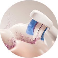 Zbliżenie na główkę szczoteczki sonicznej z niebieskimi włóknami, które czyszczą powierzchnię zębów. Wokół widać unoszące się drobinki pasty do zębów.