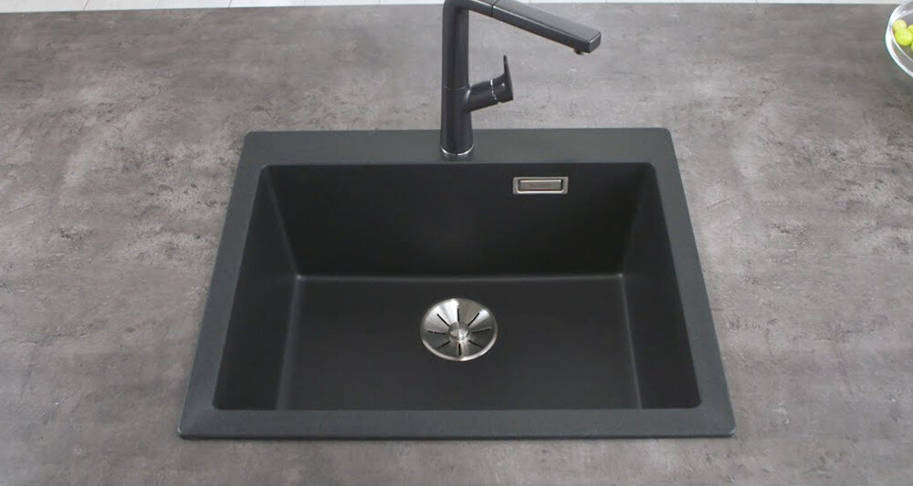 Na obrazie widoczny jest antracytowy zlewozmywak z prostokątną komorą i nowoczesną baterią. Zlewozmywak jest zamontowany w blacie o szarej, kamiennej fakturze.