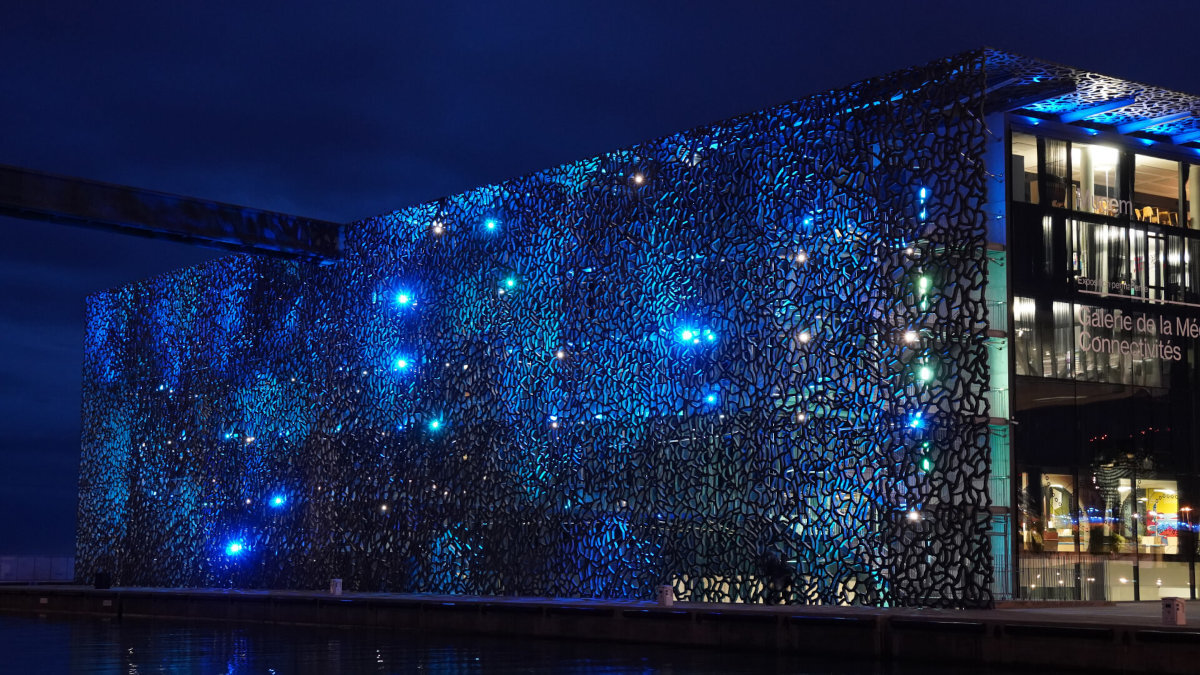 Nowoczesny budynek z ażurową fasadą oświetloną niebieskimi światłami. Na szklanej części widoczny jest napis \'Galerie de la Méditerranée\'.
