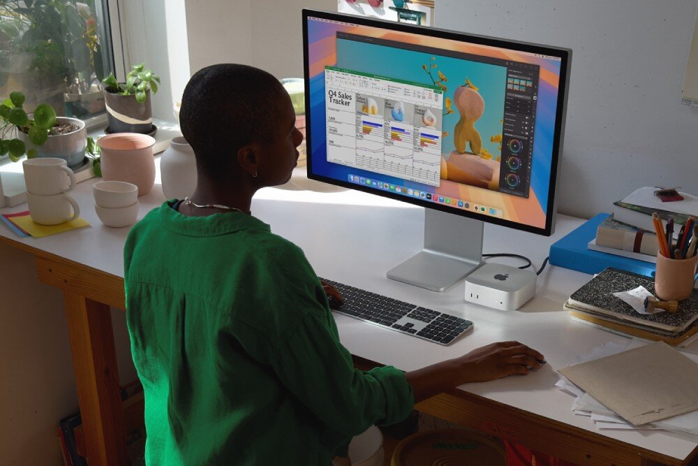 Osoba siedzi przy biurku, używając komputera z dużym monitorem, klawiaturą i myszką. Na biurku znajduje się komputer Apple Mac Mini oraz kilka roślin i książek.
