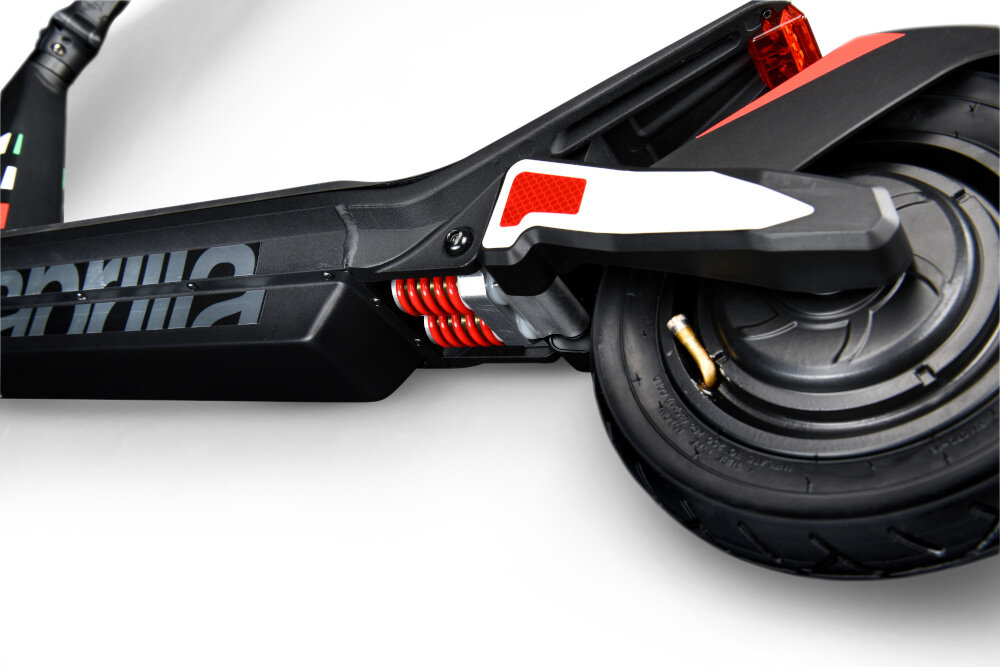 Zbliżenie na tylne koło hulajnogi elektrycznej z widocznym amortyzatorem w kolorze czerwonym. Na ramie widoczny jest napis \'aprilia\'.