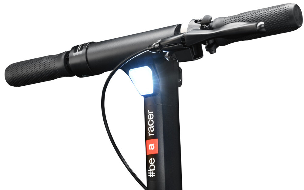 Czarny uchwyt kierownicy hulajnogi z widocznym reflektorem i napisem \'#be a racer\' na pionowej części kierownicy.