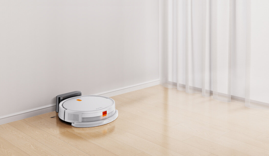 Na podłodze znajduje się biały robot sprzątający przy stacji dokującej. W tle widać zasłonę i ścianę.