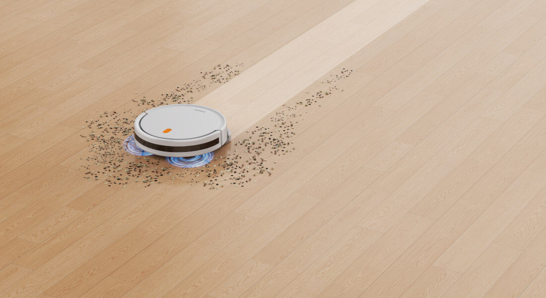 Robot sprzątający na drewnianej podłodze, zbierający rozsypane drobinki. Na urządzeniu widoczne jest logo producenta.