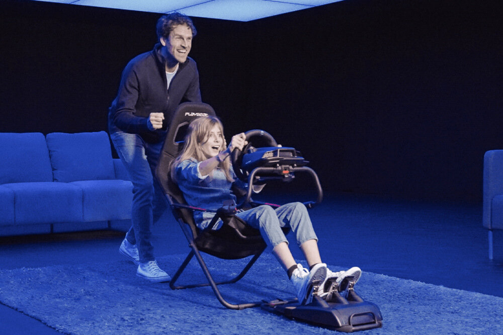 Osoba siedzi w czarnym kokpicie PLAYSEAT Challenge Actifit, trzymając kierownicę symulatora wyścigowego. W tle widoczna jest kanapa i osoba stojąca obok, a cała scena jest oświetlona niebieskim światłem.